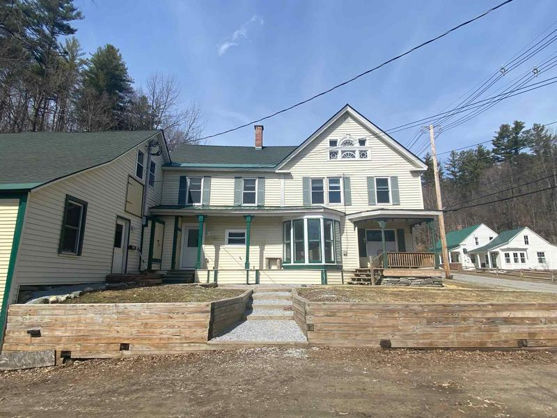 The exterior of a two-story house features a light-colored facade with green trim. A spacious porch with seating is positioned at the front, and a gravel pathway leads to the entrance. Surrounding greenery and nearby homes enhance the quaint atmosphere.