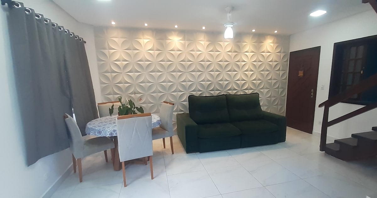A living area is seen with a green sofa and a dining set comprising four upholstered chairs surrounding a circular table. A textured wall with triangular designs adds depth to the space, complemented by soft lighting from recessed ceiling fixtures. A ceiling fan enhances air circulation.