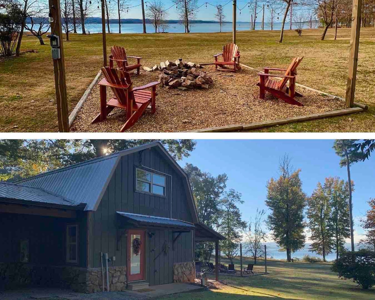 A fire pit is surrounded by four red adirondack chairs on a grassy area, with a view of the lake in the background. The cabin exterior features a rustic design, with a stone foundation and large windows reflecting the surrounding trees.