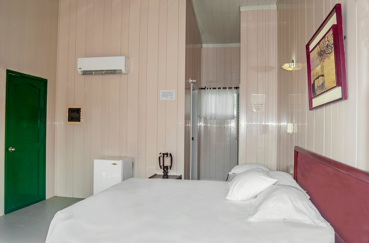 A spacious sleeping area features a large bed with several pillows. A small refrigerator and an air conditioning unit are visible. A bathroom area is partially seen at the back, separated by an open doorway. The walls are clad in light-colored wood paneling.