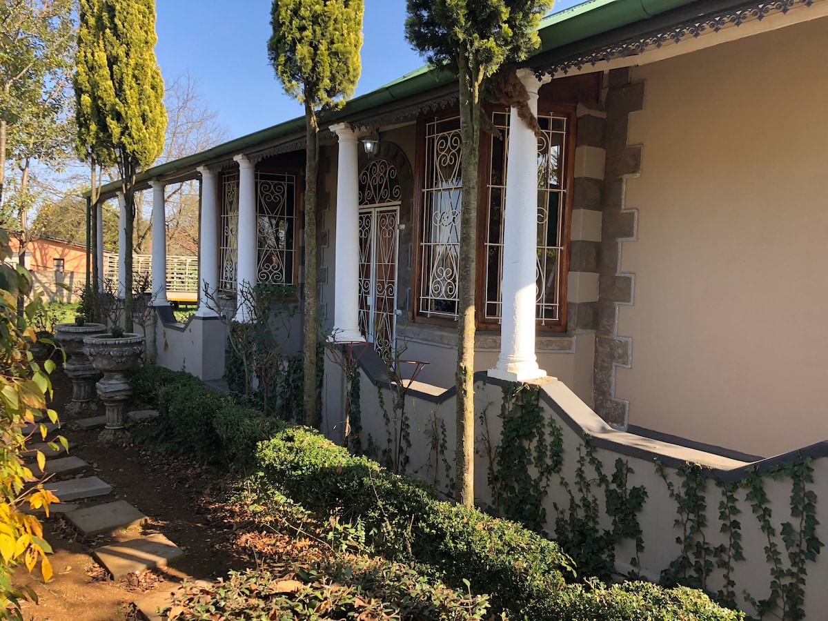 The exterior of the house features tall columns supporting a covered veranda, surrounded by neatly trimmed hedges and climbing greenery. The walls exhibit a combination of stones and plaster, creating a characterful appearance. A pathway leads toward the entrance, enhancing the welcoming feel of the space.