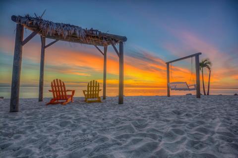 A serene beach scene is captured at sunset, featuring two brightly colored Adirondack chairs positioned on soft white sand. A swing hangs from a wooden frame nearby, with gentle waves lapping at the shore and a vibrant sky illuminated in hues of orange and pink.