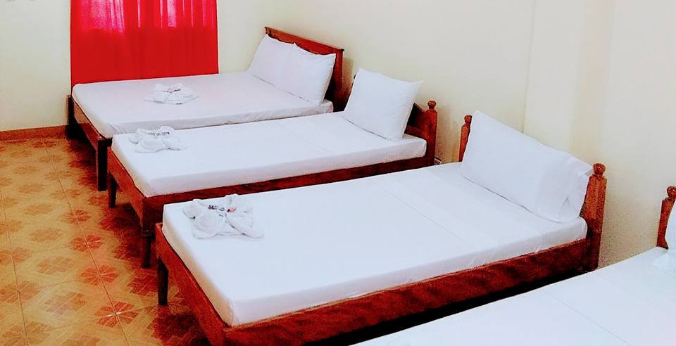 The room features four wooden beds arranged neatly, each dressed in crisp white linens. Plush pillows are placed at the head of each bed, while folded towels are neatly positioned on top. Natural light enters through a red-curtained window, providing a warm hue.