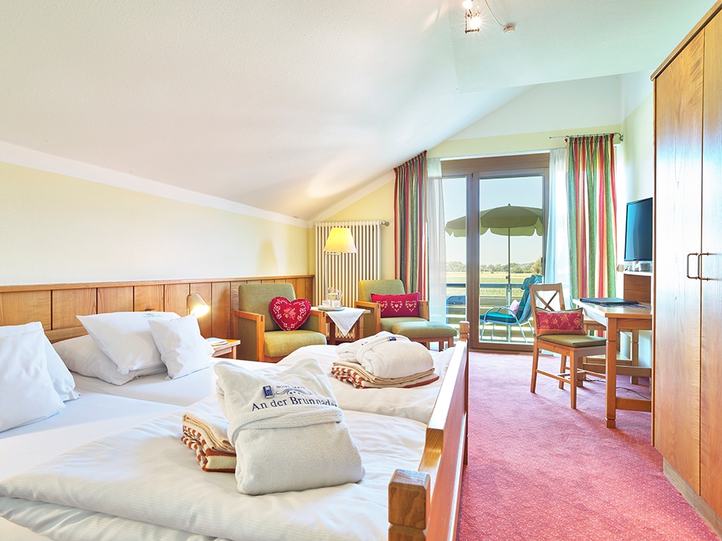 A bright room features two spacious beds with soft linens and stacked towels. A cozy seating area includes a green sofa and a small table. A balcony door leads to outdoor seating under an umbrella, with neighboring greenery visible through the glass.