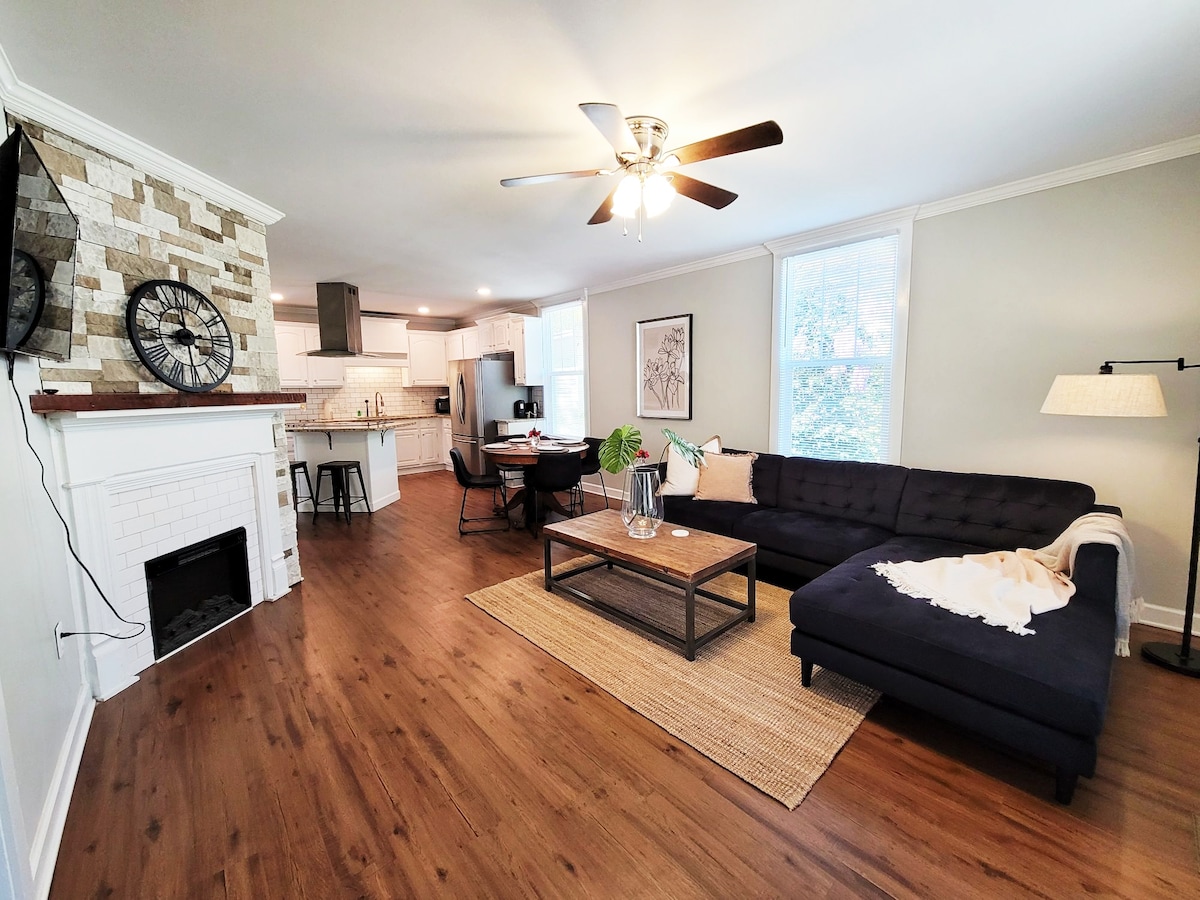 A welcoming living space features a dark sectional sofa with a light throw, arranged around a rectangular wooden coffee table. Light wood flooring enhances the area, complemented by a textured area rug. A modern ceiling fan and large windows contribute to the bright atmosphere.