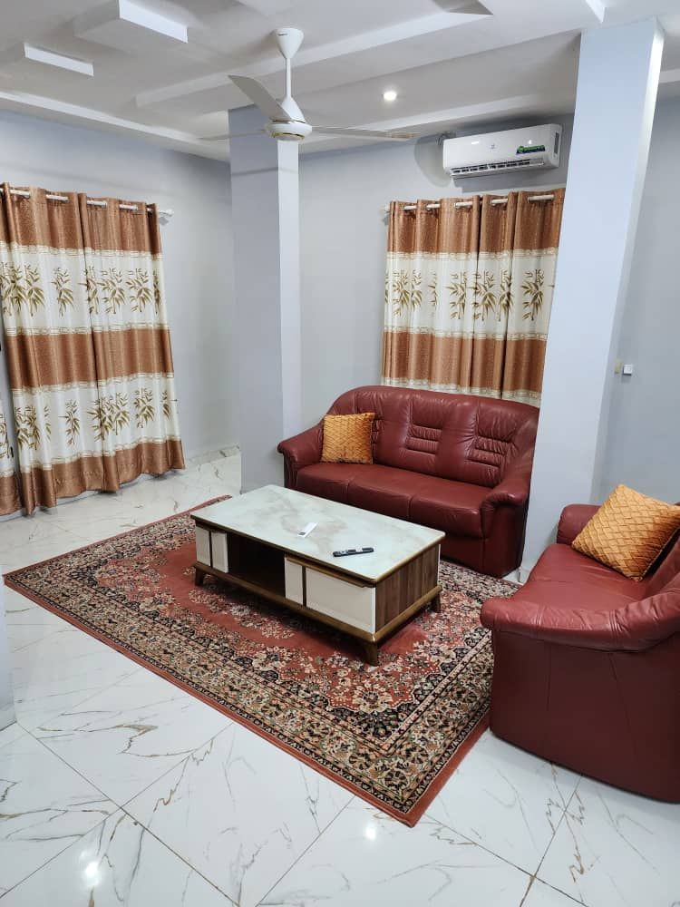 A living area is furnished with a pair of red leather sofas and an elegant coffee table at the center. A decorative rug covers the tiled floor, while floor-to-ceiling curtains in earthy tones provide privacy and add warmth to the space.