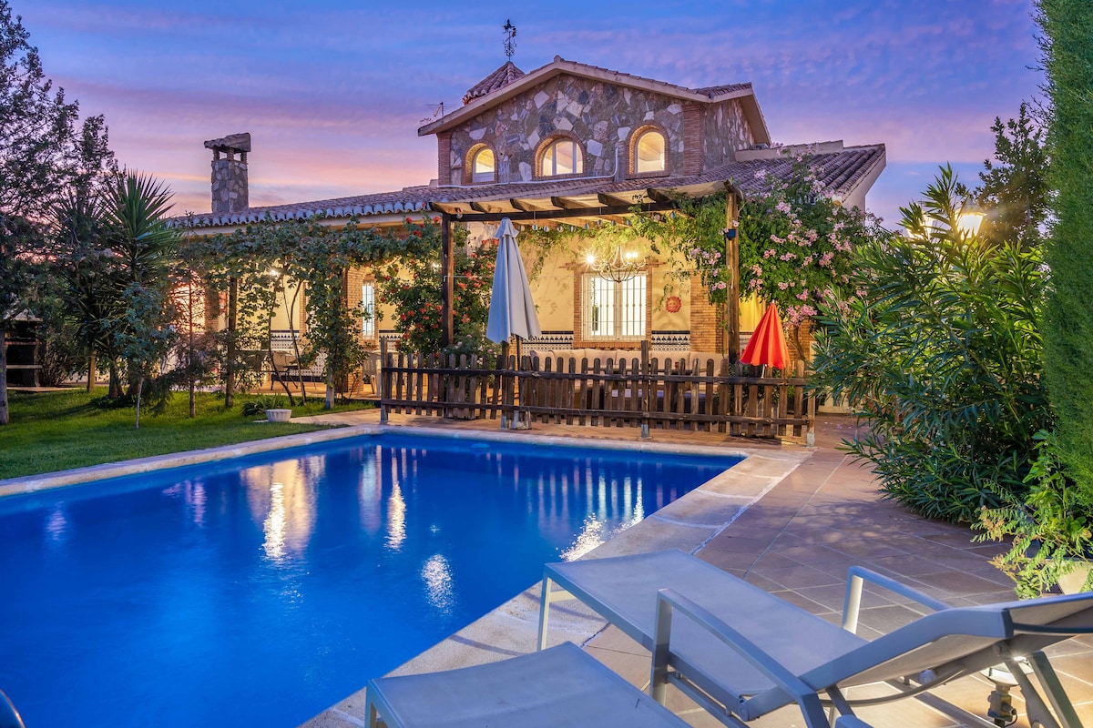 A tranquil outdoor scene features a well-maintained pool surrounded by lounge chairs. A charming stone house with decorative vines is illuminated by twilight. Colorful umbrellas provide shade for the deck area, while lush greenery enhances the serene setting.