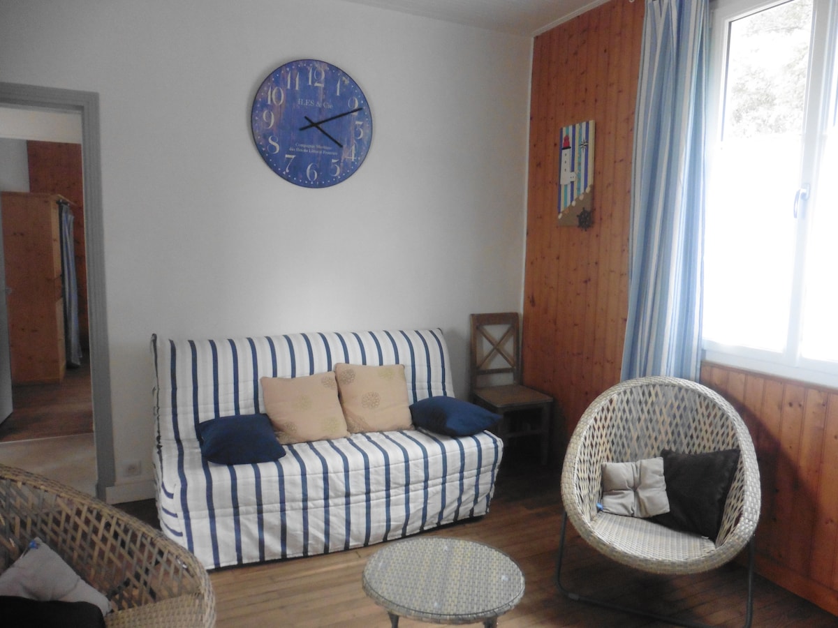 A living area features a striped sofa with decorative pillows, accompanied by two woven chairs. A light wooden table is centered in the space, while a large wall clock adds an element of timing. Natural light filters through the nearby window.