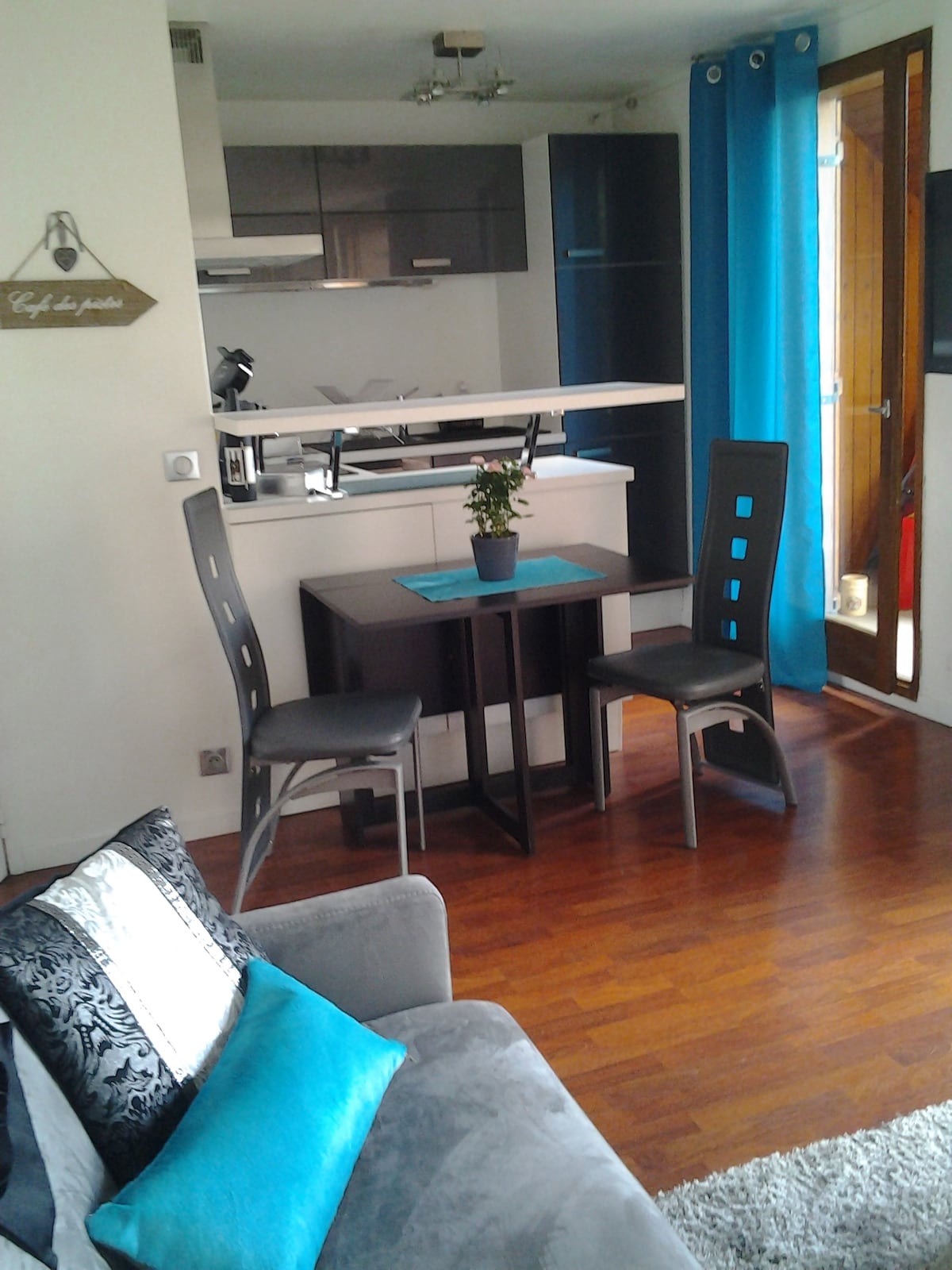 A modern living area features a compact dining table with two chairs placed on a rich wooden floor. An open kitchen is visible in the background, complemented by sleek cabinetry. Light blue curtains frame the doorway, offering a touch of color, while a small plant adds a hint of greenery.