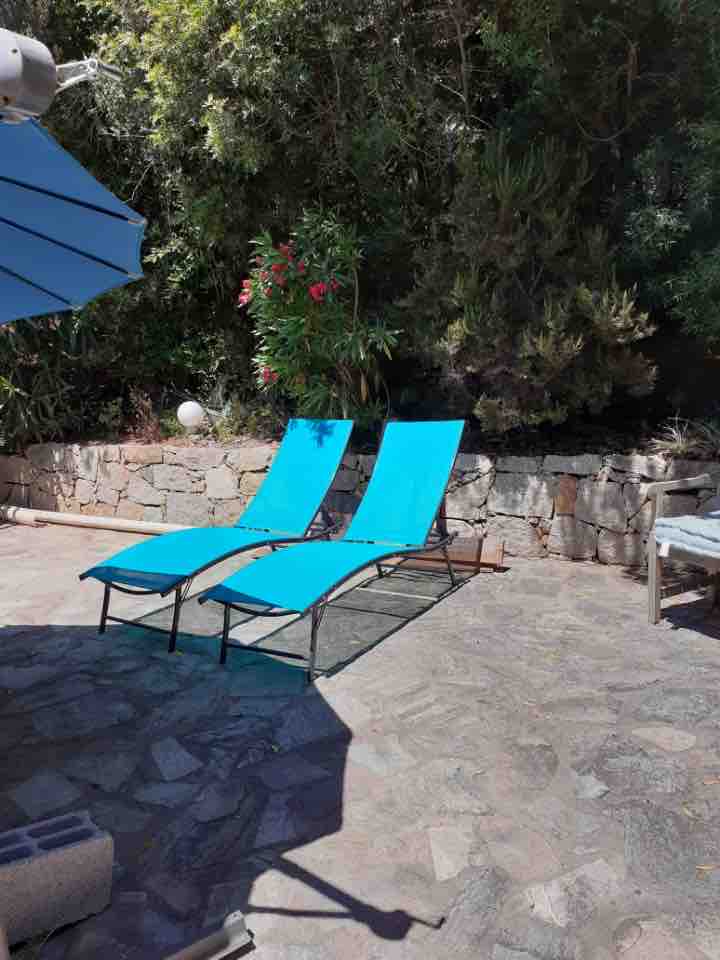 Two blue lounge chairs are positioned on a stone patio, surrounded by lush greenery. A large umbrella provides shade from the sun, while a stone wall adds privacy to the space.