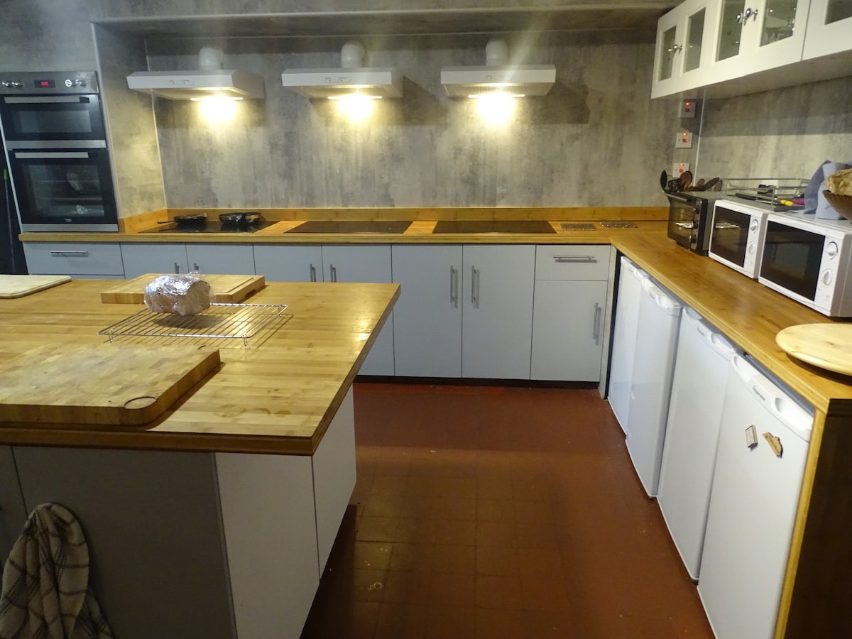 A well-equipped kitchen features light-coloured cabinetry and a spacious wooden countertop. Multiple cooking appliances are visible, including ovens and microwaves. Adequate lighting is provided by overhead fixtures, and a cooling area with white refrigerators is located along one side.
