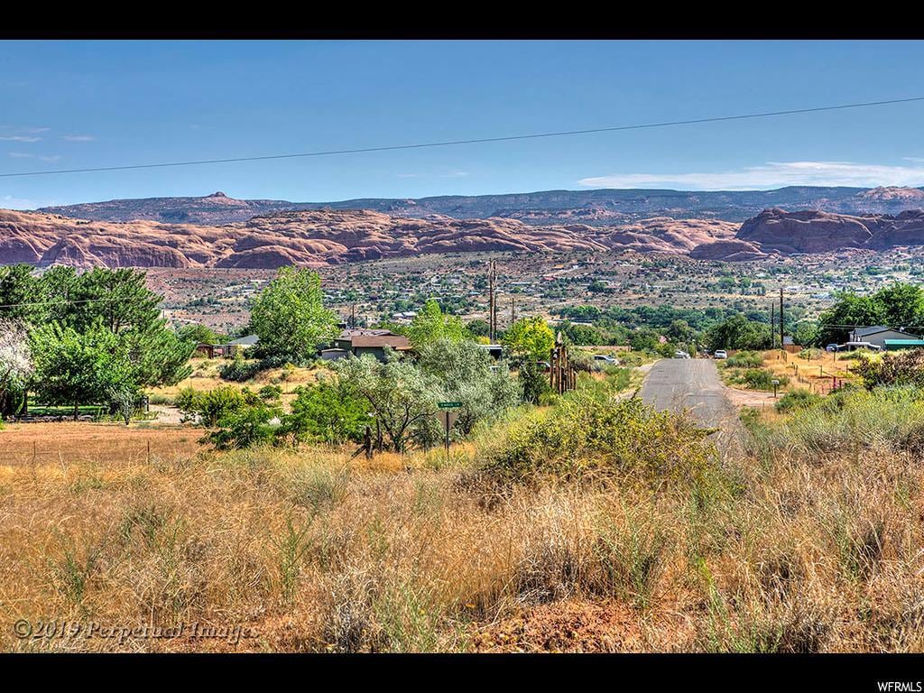 A scenic view captures the surrounding landscape, featuring rolling hills and vibrant greenery beneath a clear blue sky. The road leads towards the horizon, where striking red rock formations rise against the backdrop of distant mountains.