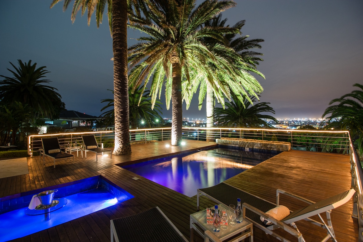 An outdoor space is illuminated at night, featuring a large swimming pool and a separate jacuzzi. Surrounding palm trees provide a tropical feel, while wooden decking enhances the area. Comfortable lounge chairs are positioned for relaxation, and subtle lighting highlights the serene atmosphere.