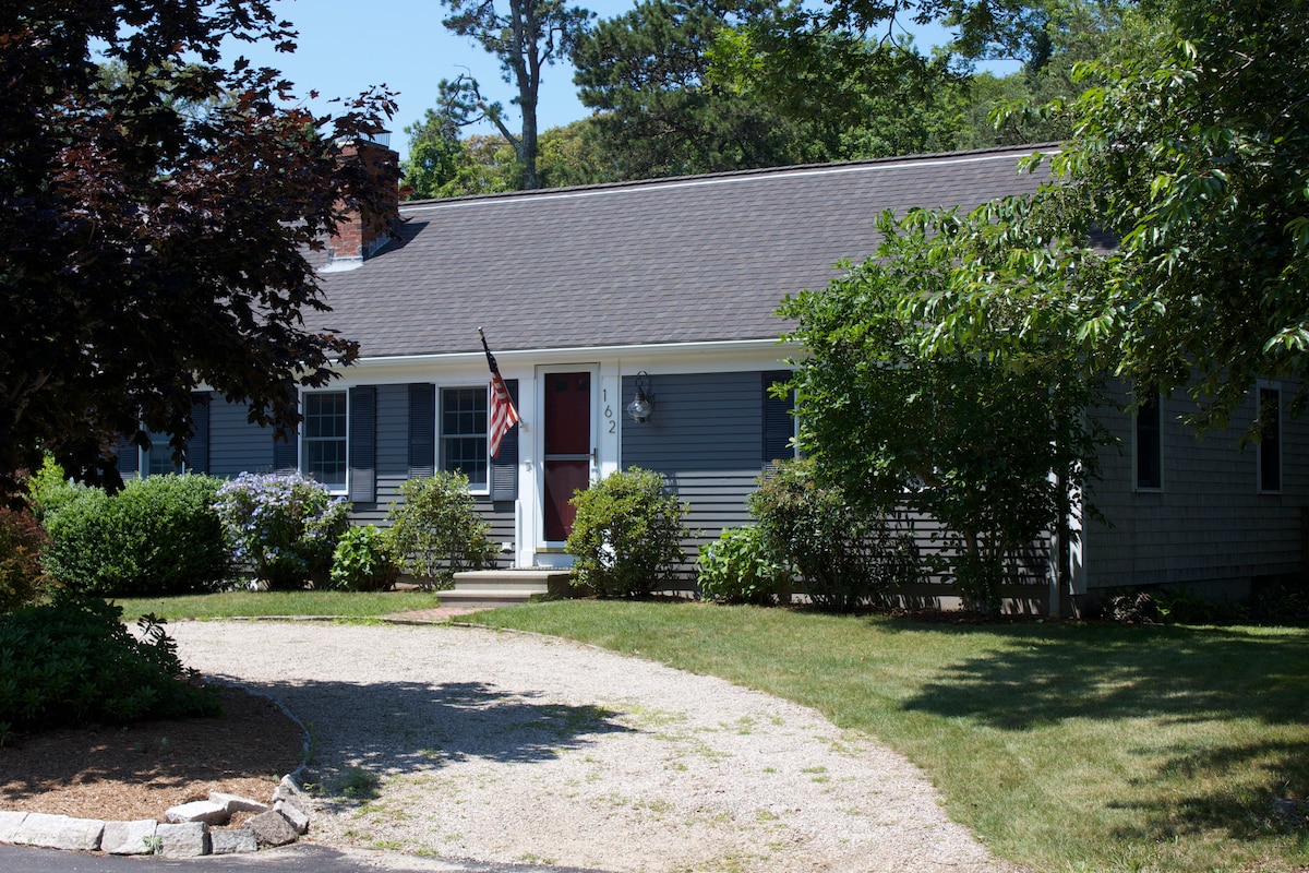 A single-story ranch-style home is shown, featuring grey siding and white trim. A welcoming front porch is framed by lush greenery and flowering shrubs. A gravel driveway leads to the entrance, complemented by well-maintained landscaping and an American flag displayed by the door.