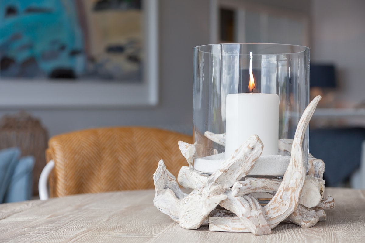 A glass candle holder features a white candle surrounded by a natural driftwood base, adding a coastal touch to the decor. Soft light from the candle illuminates the table, with a blurred background of contemporary furnishings and artwork.