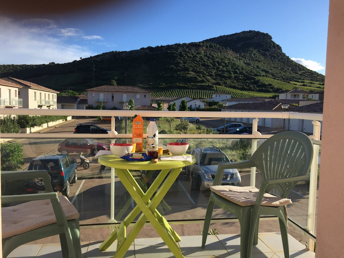 A small balcony features a bright green table and two light-colored chairs, set against a backdrop of rolling hills. The table is laid with bowls and drinks, while the view includes surrounding buildings and parked cars.