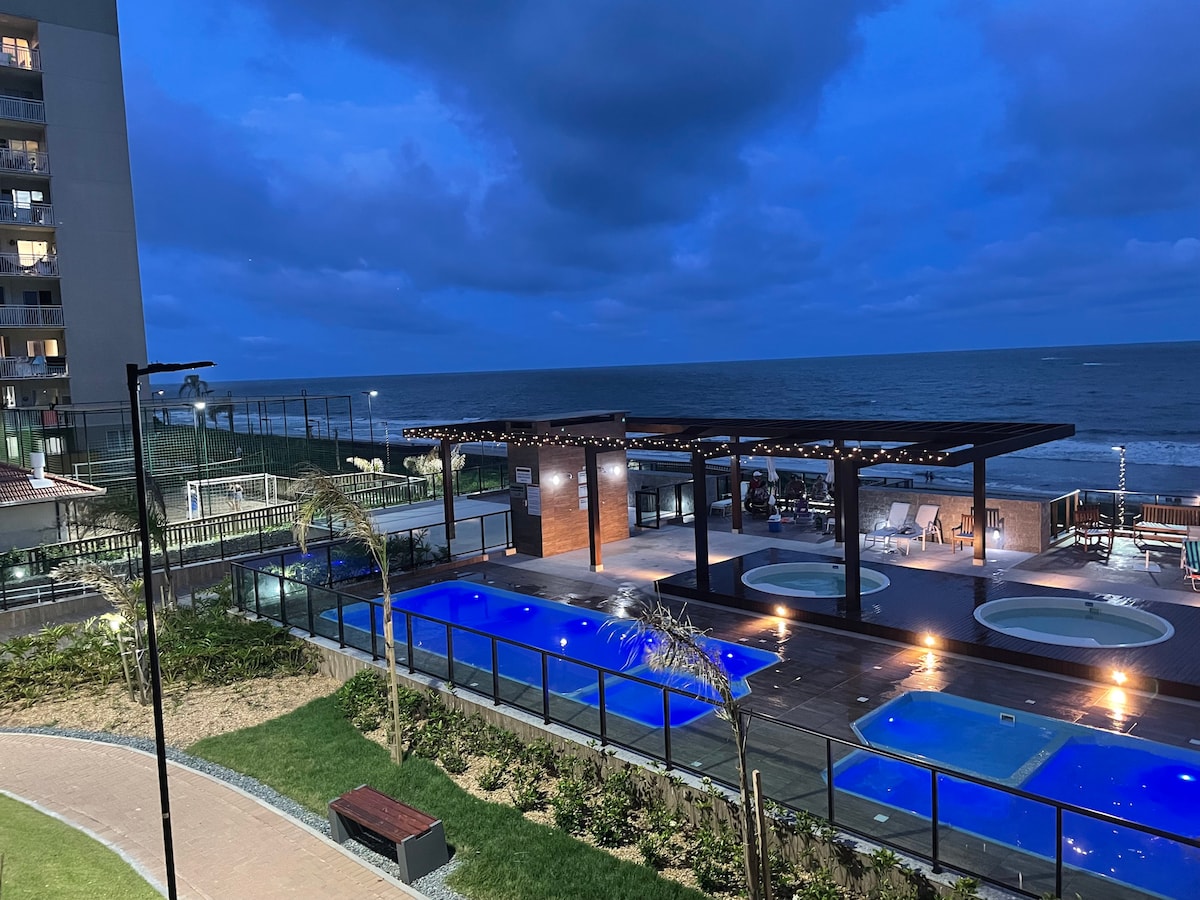 An evening view showcases the outdoor pool area illuminated by soft lights, with two pools set against the backdrop of the ocean. Lounge chairs and a shaded seating area add to the inviting atmosphere, framed by well-manicured greenery and pathways.