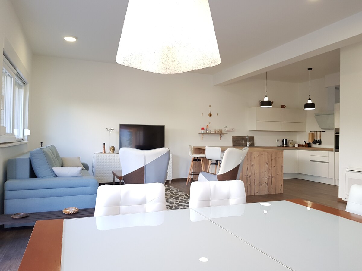 A bright living area reveals a comfortable blue sofa next to a modern television. The dining table reflects natural light, accompanied by white chairs. A kitchen area features a wooden counter and contemporary lighting, emphasizing an open and inviting layout.