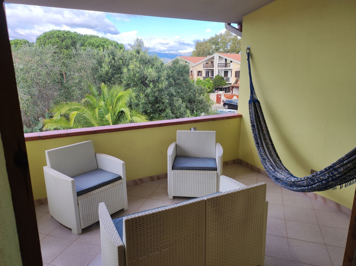 A balcony area is furnished with a set of four white wicker chairs with blue cushions and a hanging hammock. Foliage is visible in the background, providing a natural setting. The space is designed for relaxation with ample room for seating.