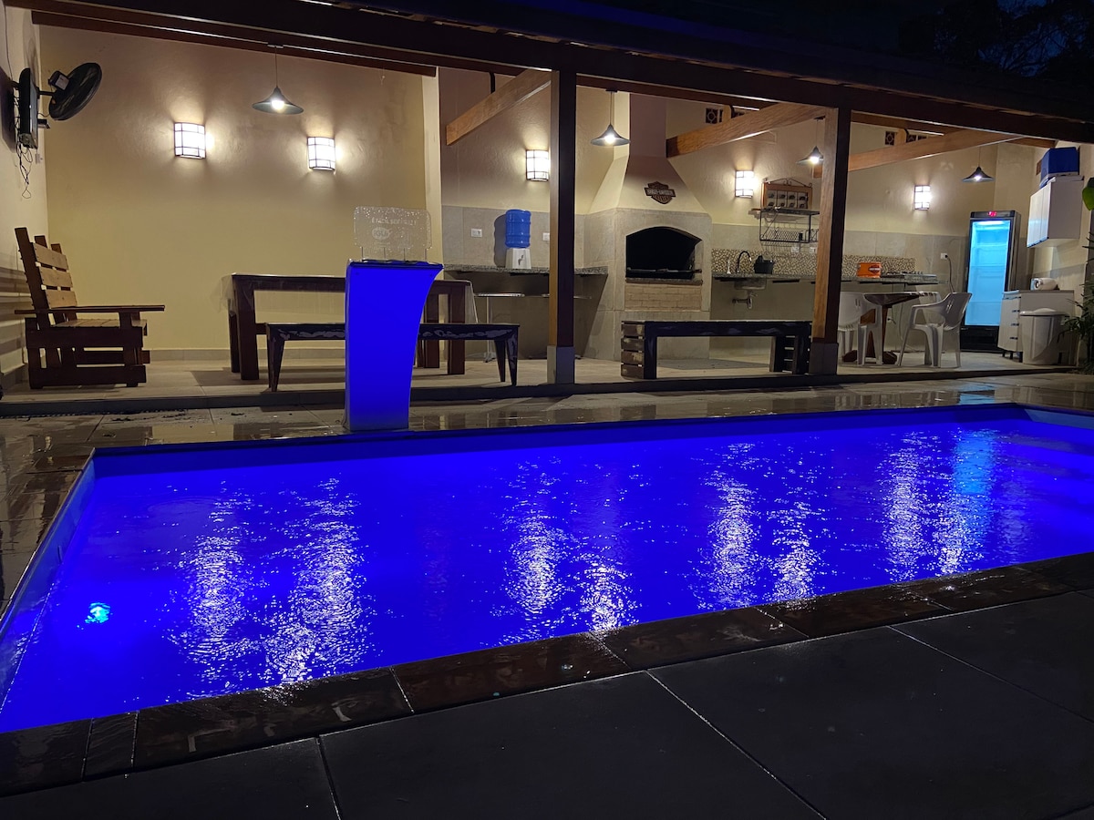 An inviting outdoor space is showcased, featuring a swimming pool with illuminated blue waters. The area includes a covered barbecue section with seating, and a wood-burning oven, creating a functional and comfortable gathering spot.