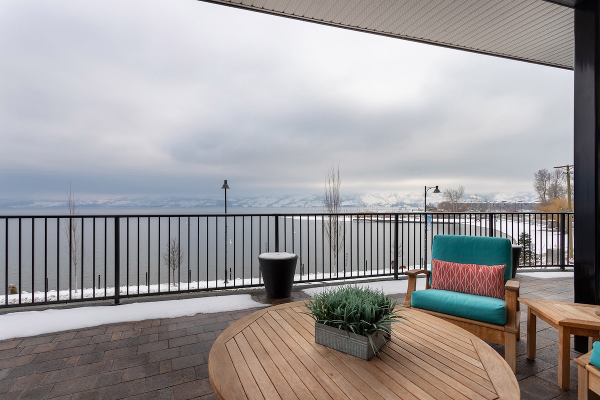 A spacious patio features a round wooden table surrounded by comfortable seating, offering a view of the calm lake and distant mountains. The area is enhanced by potted plants and outdoor lighting, with snow visible on the ground and an overcast sky in the background.