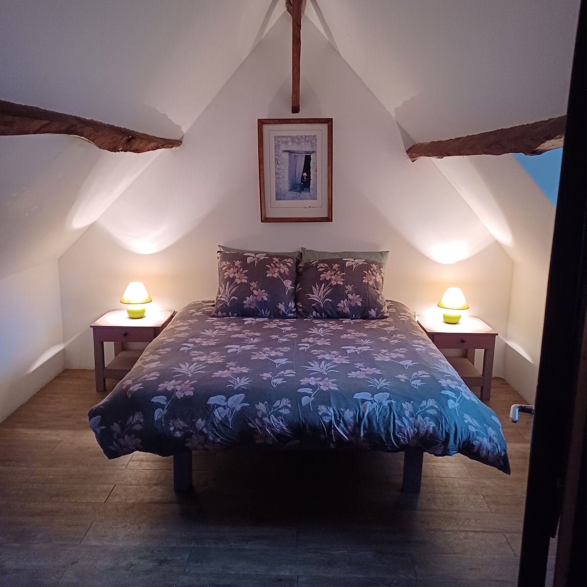 An inviting bedroom features a spacious bed adorned with floral bedding. Soft lighting from two bedside lamps creates a calming ambiance, highlighting the wooden beams on the sloped ceiling. A framed picture decorates the wall above the bed, contributing to the serene atmosphere.