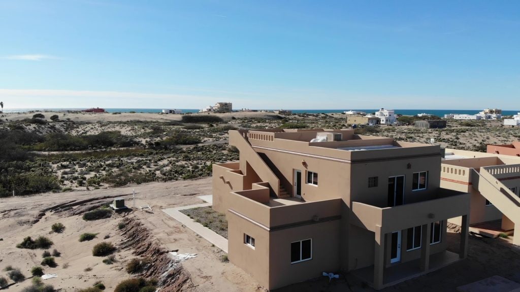An aerial view of a two-story beach home is presented, showcasing its sandy surroundings and proximity to the ocean. The exterior features light-colored walls, multiple windows, and a staircase leading to the rooftop terrace, ideal for enjoying expansive views of the landscape.