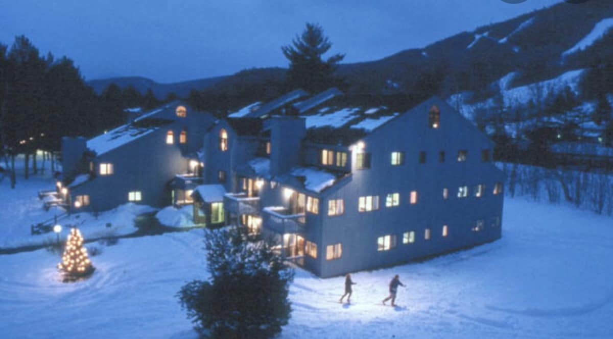 A multi-level building with a blue exterior is surrounded by snow-covered terrain. Warm yellow lights illuminate the windows, contrasting against the twilight sky. In the foreground, two individuals ski along a snowy path, while a decorated tree adds a festive element to the winter scene.