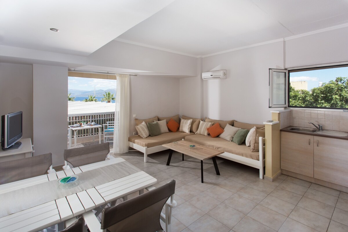 A spacious living area features a comfortable sectional sofa adorned with colorful cushions. A dining table with grey chairs is positioned nearby. Natural light enters through large windows, showcasing views of the outdoors. A compact kitchen area is visible in the corner.