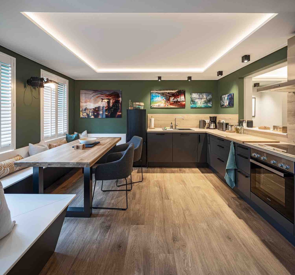 A spacious kitchen and dining area features modern appliances and sleek cabinetry. A large wooden dining table is surrounded by comfortable chairs. Colorful artwork adorns the walls, while natural light enters through multiple windows, enhancing the contemporary design.