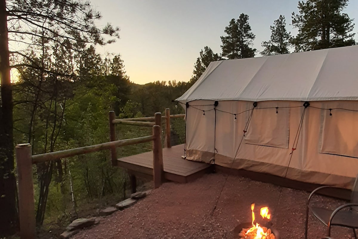 A spacious tent is positioned on a wooden platform surrounded by trees, with a small fire pit in the foreground. The setting sun casts a warm glow, creating a peaceful atmosphere in the natural landscape.