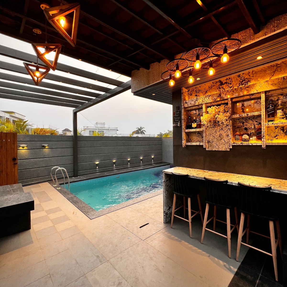 An outdoor space features a sleek pool surrounded by tiled flooring. A bar area with high stools is visible, showcasing decorative shelves and ambient lighting. The space is enclosed by stylish walls, providing a sense of privacy and comfort, complemented by palm trees in the background.