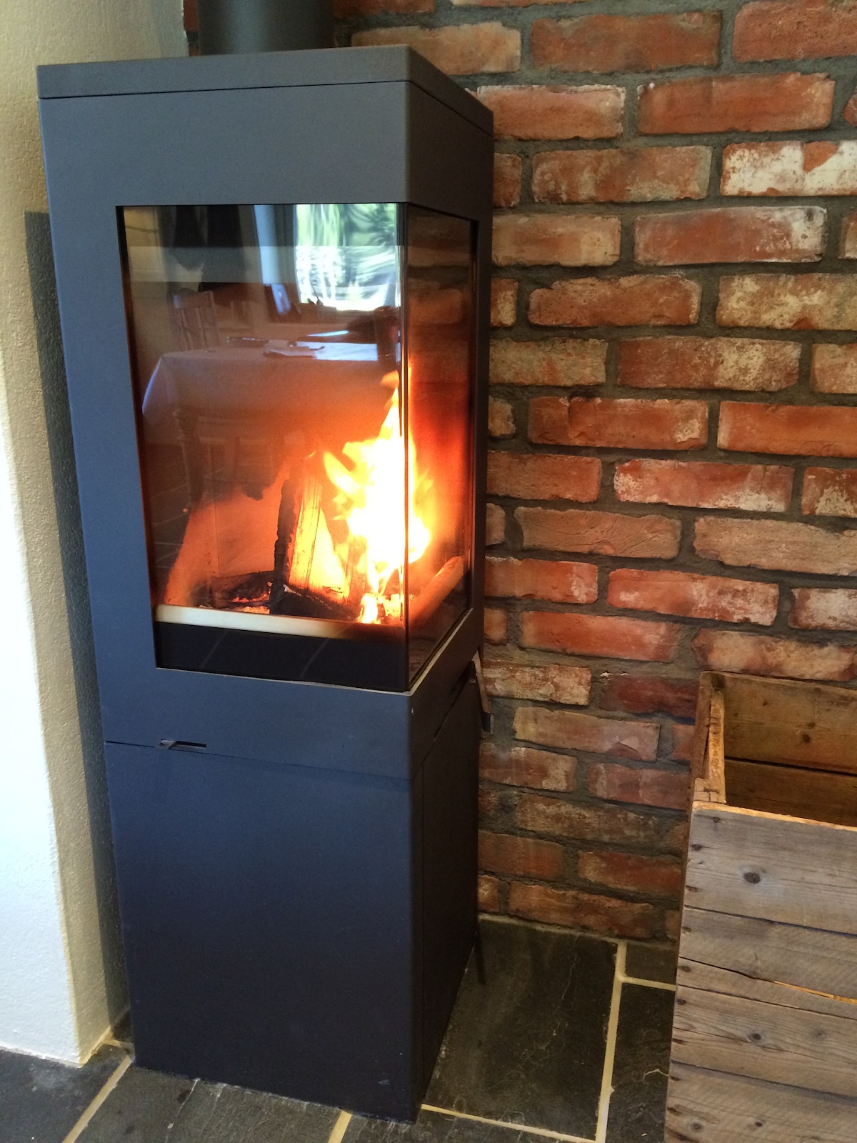 A modern wood-burning stove is positioned against a rustic brick wall, providing warmth with a visible flame inside. The surrounding area features dark stone flooring and a wooden crate is placed nearby, adding to the cozy ambiance.