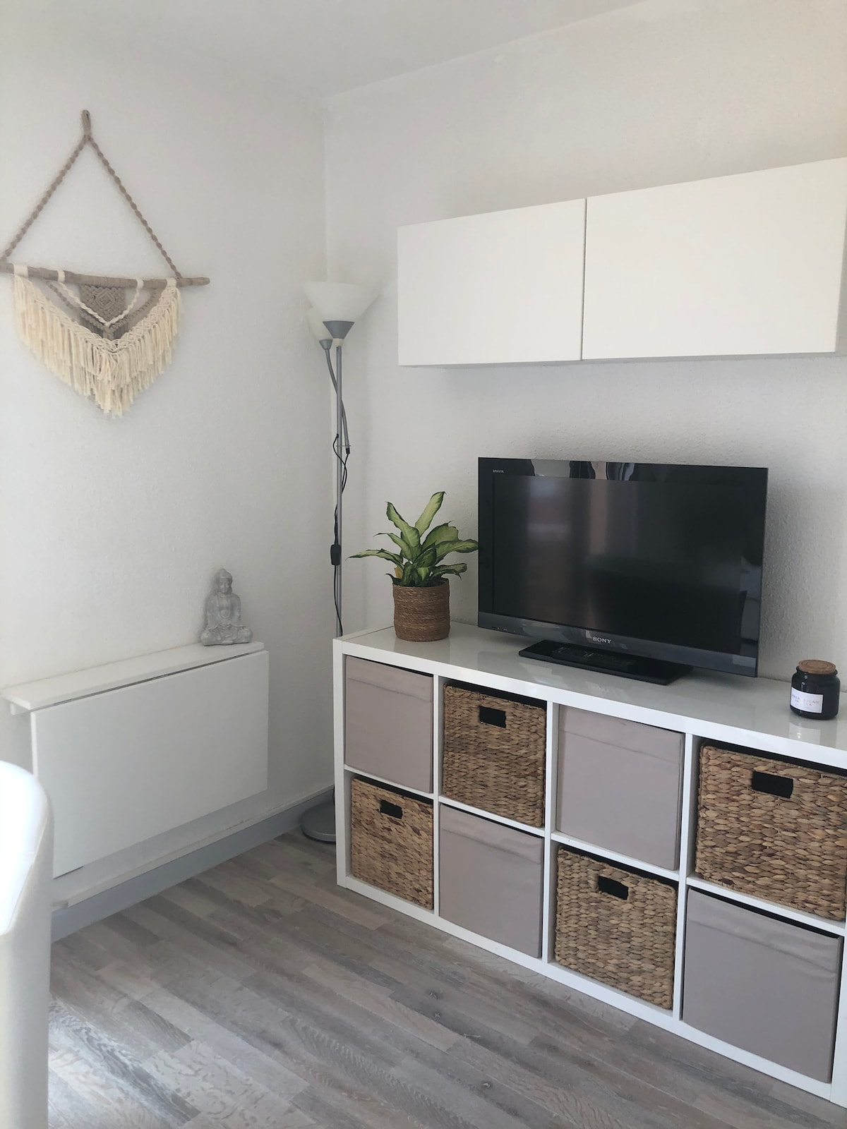 A living area features a minimalist design with light-colored walls and wooden flooring. A television is mounted on a white cabinet, accompanied by a potted plant. Beneath the TV, storage boxes made of woven natural materials are arranged, providing a tidy appearance.