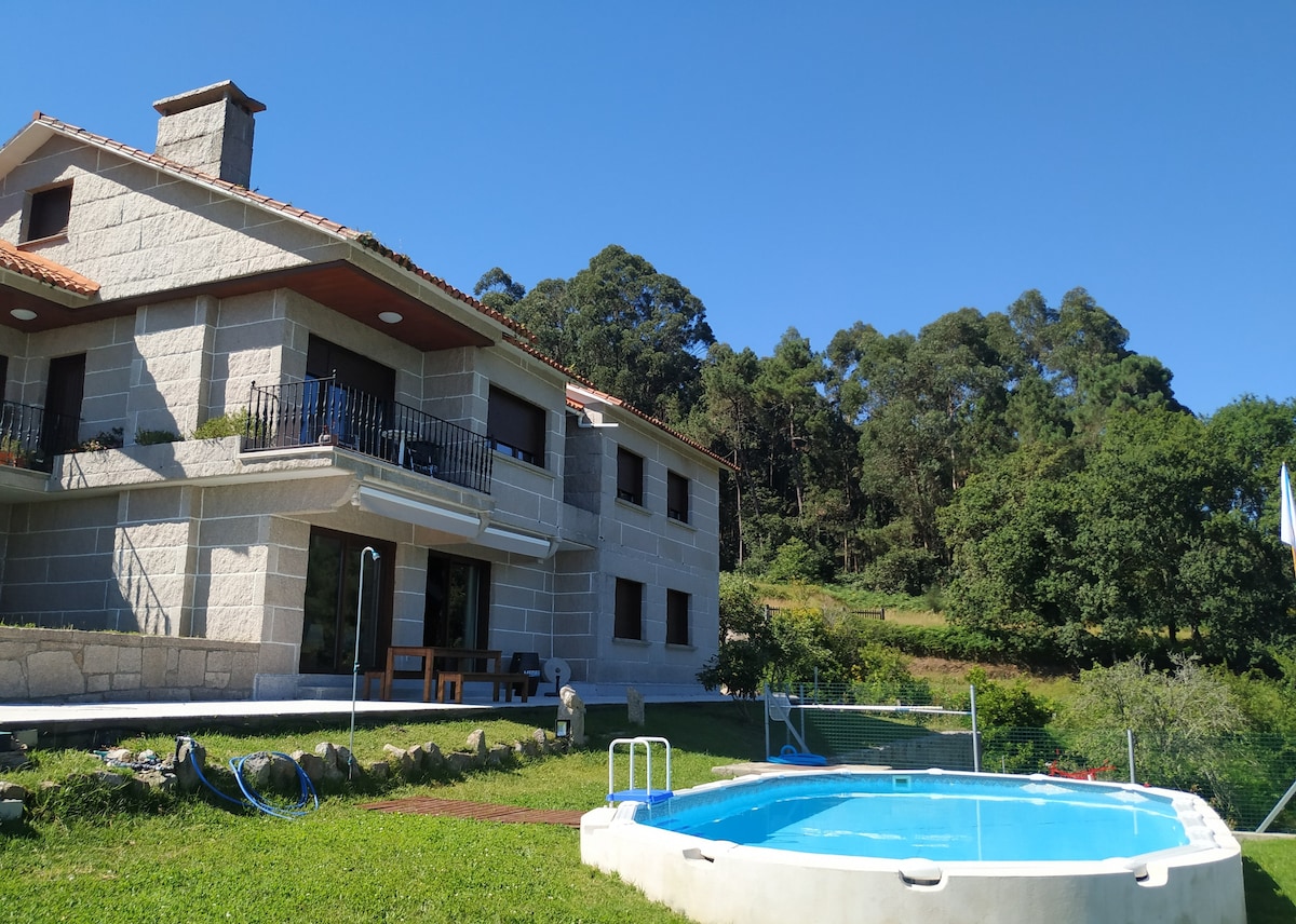 A stone-clad house sits amidst greenery, featuring a small circular pool beside a well-kept lawn. The building's two stories showcase a blend of modern architecture and natural surroundings, with trees and hills visible in the background under a clear blue sky.
