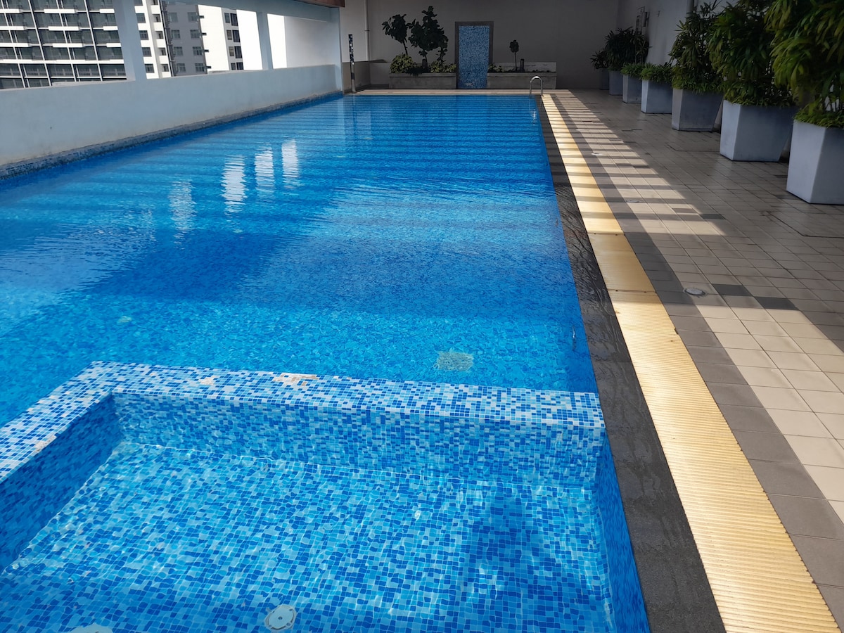 A serene rooftop pool features clear, turquoise water, harmoniously reflecting light. Surrounding potted plants create a calming atmosphere, while the pool deck offers ample space for relaxation. The area is illuminated by a soft golden edge, complementing the crispness of the water tiles.