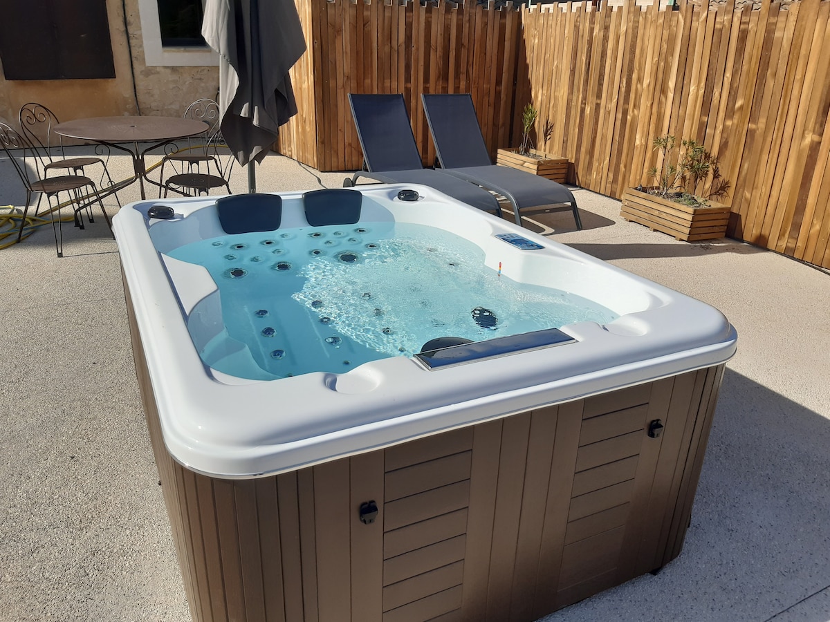 A private hot tub is positioned outdoors, surrounded by a wooden fence. Two lounge chairs are placed nearby under a gray umbrella, with a small table visible in the background. The hot tub features a sparkling blue water surface, inviting relaxation.
