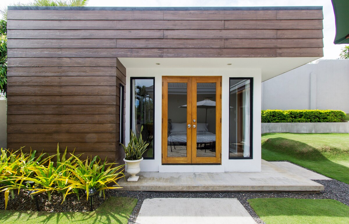 The modern guest house exterior features a sleek wooden facade and large glass doors leading to an outdoor patio. Lush green landscaping, including potted plants, complements the entryway, which is set against a well-maintained lawn.