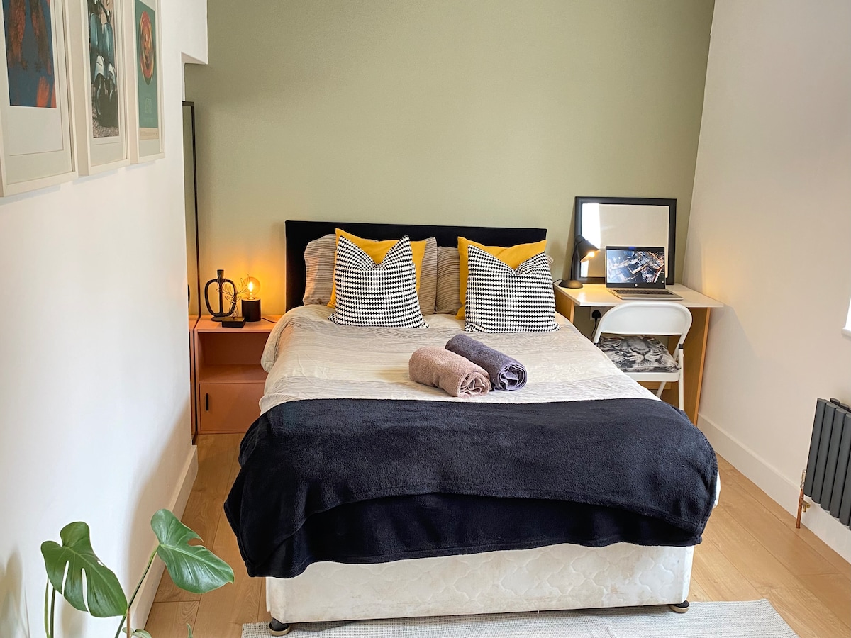 A well-appointed double bedroom features a spacious bed topped with contrasting yellow and patterned cushions. Soft towels are neatly arranged, and a bedside table displays an ambient lamp. A small white console provides extra storage. Light-colored walls enhance the room's brightness.