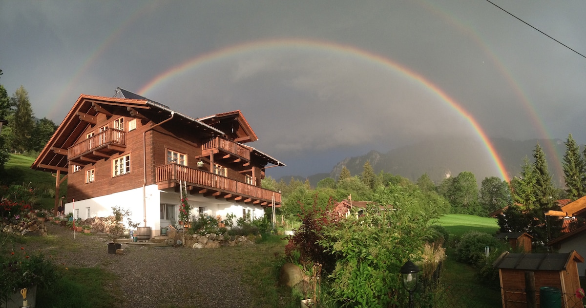 A charming wooden building is set against a backdrop of lush greenery, with a vibrant double rainbow arching across the sky. The residence features balconies and a well-maintained garden area, enhancing the peaceful rural landscape.
