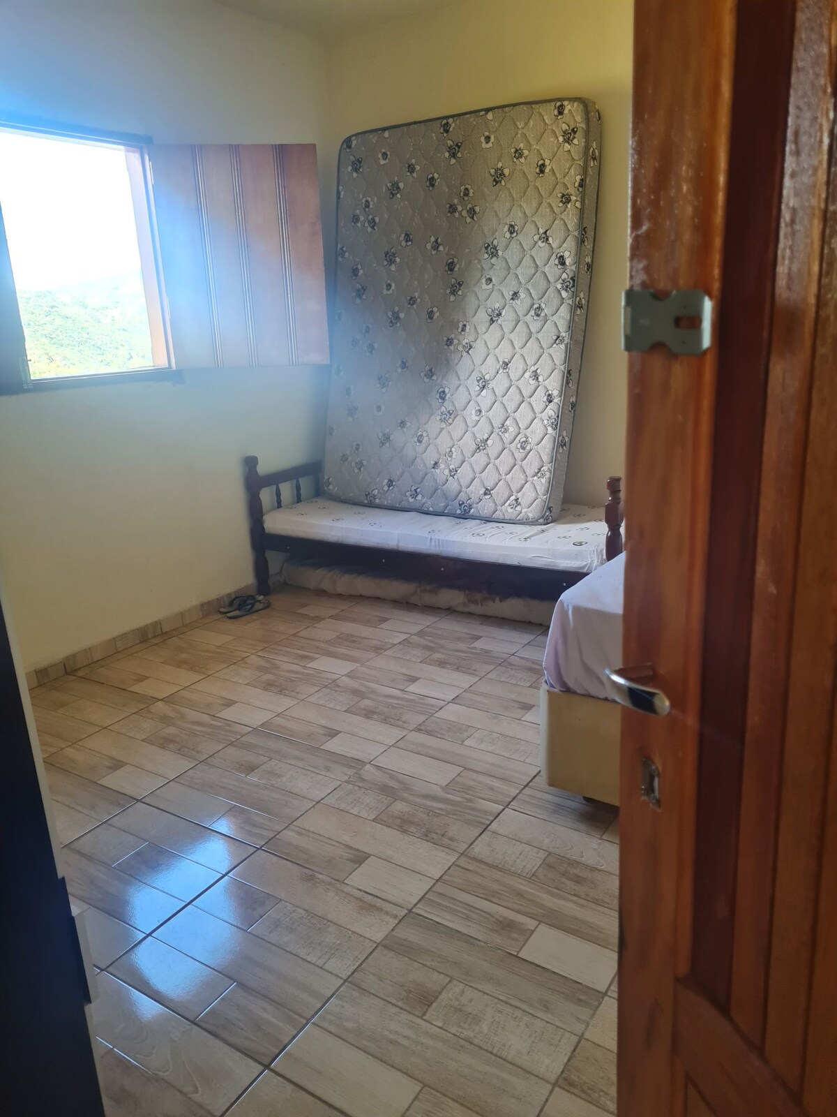 A simple room features a large window that allows natural light to fill the space. A mattress leans against the wall, and a small bench is positioned beside it. The tiled floor provides a clean and functional surface.