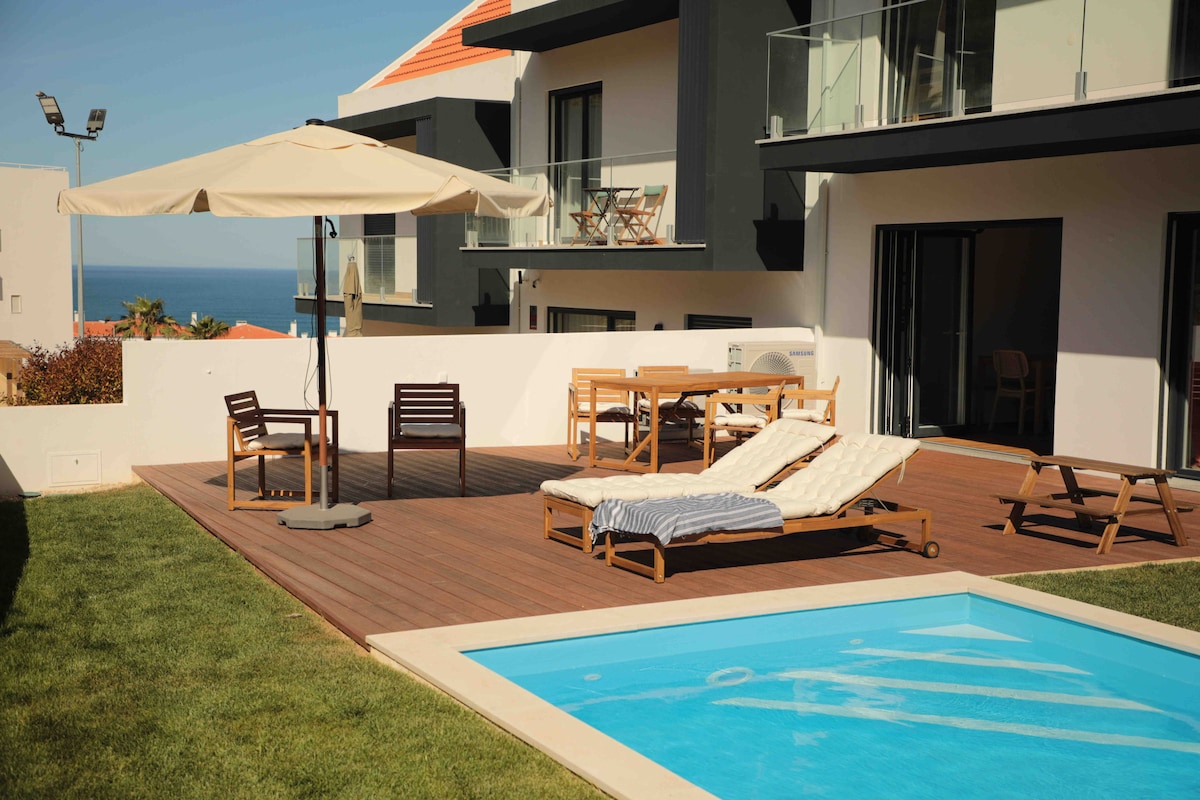 The outdoor area showcases a private terrace with a wooden deck, featuring a pool, sun loungers, and a dining table. An umbrella provides shade, while plush seating offers a relaxing space for enjoying views of the surrounding area.