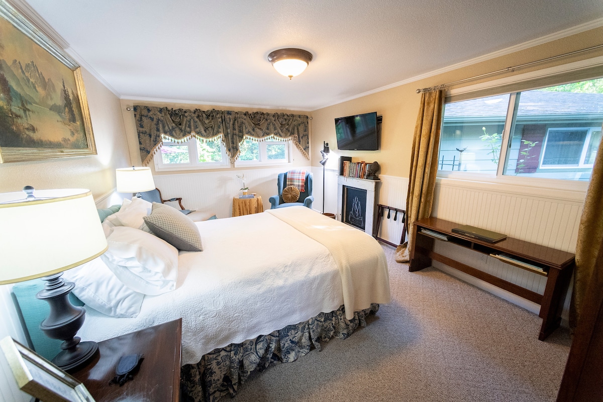 A well-appointed bedroom features a spacious bed with a decorative bedspread and an assortment of pillows. A flat screen TV is mounted on the wall, and a large window offers natural light. A small table and lamp are positioned beside the bed, contributing to a functional layout.
