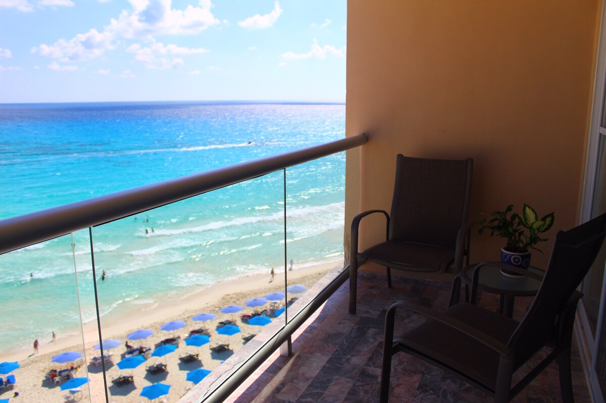 A private balcony offers views of the Caribbean Sea, with gentle waves lapping at the sandy beach below. Two comfortable chairs are positioned beside a small table, while a potted plant adds a touch of greenery. Beach umbrellas can be seen lining the shore.