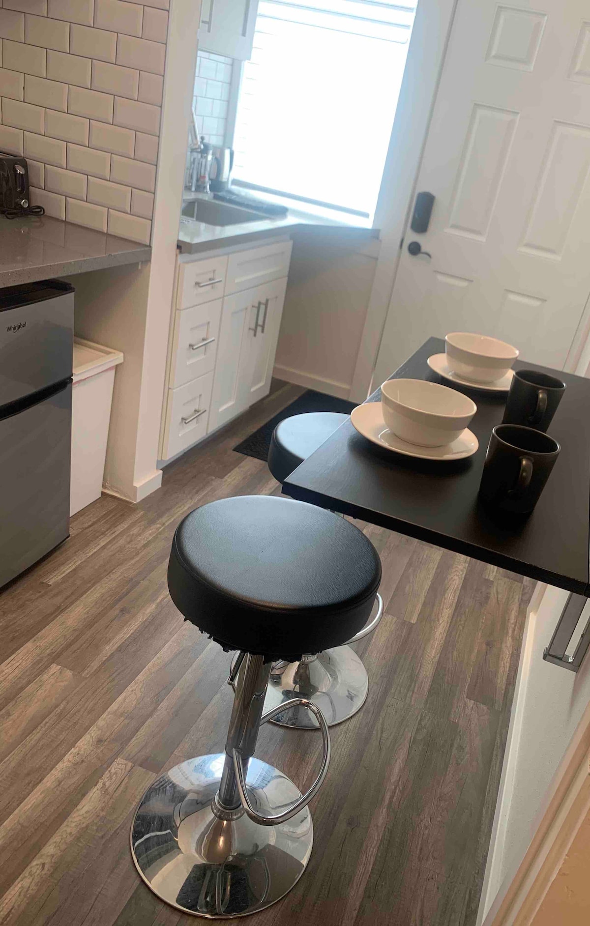 The kitchen area features modern appliances, including a compact refrigerator and a stylish countertop. A small dining bar with three stools is positioned near a window, complemented by three bowls and two mugs resting on the tabletop.