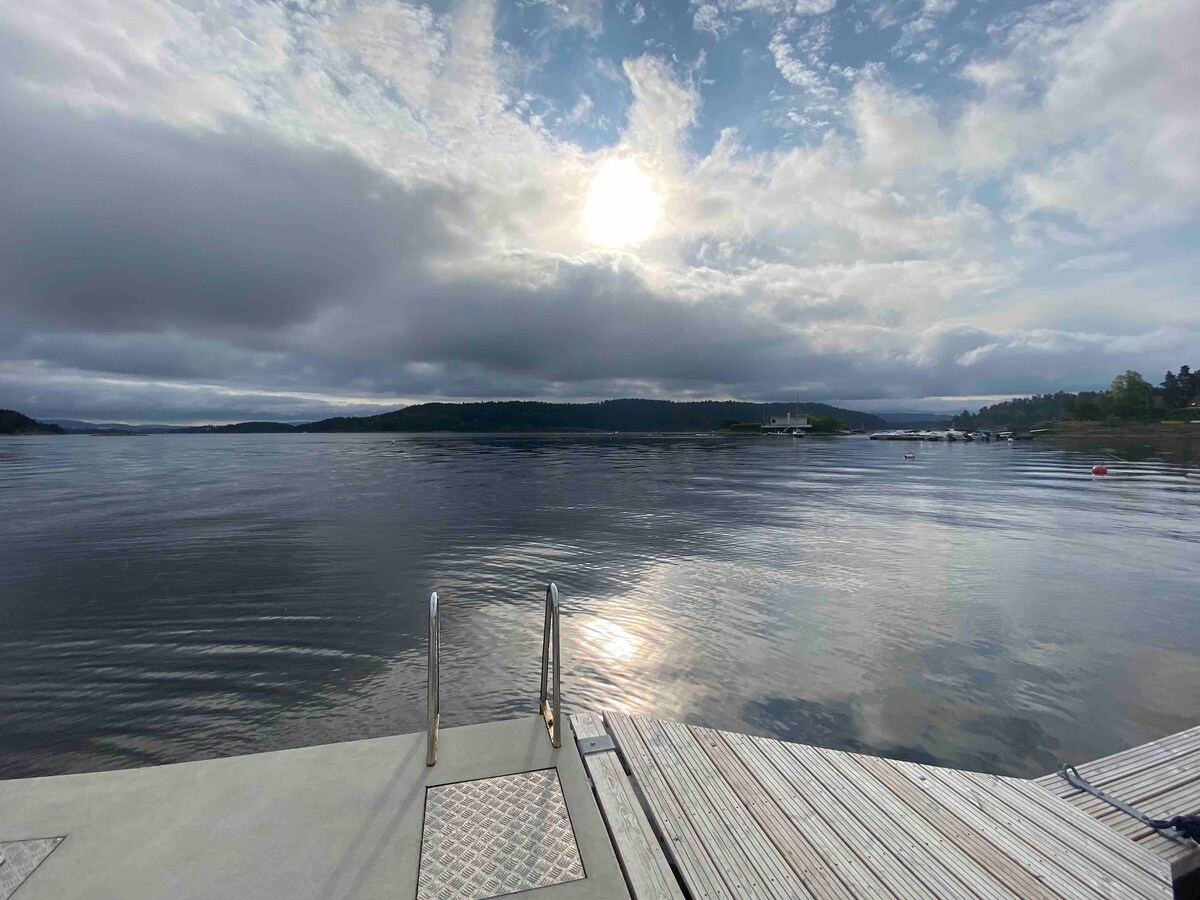 A calm waterfront scene is depicted, featuring gentle waves reflecting the sun. The dock is positioned at the forefront, with metal stairs leading to the water. A soft cloud cover enhances the serenity, while distant land is seen on the horizon.