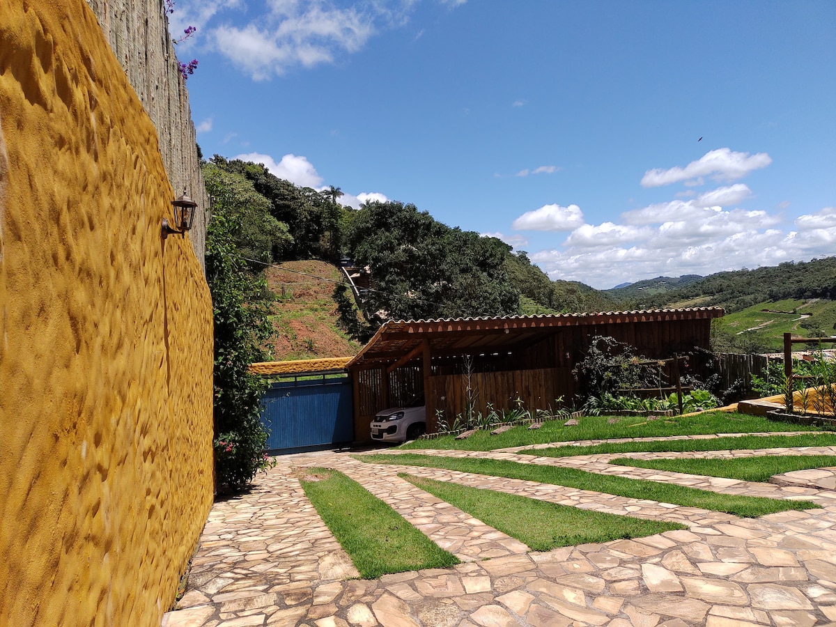 The outdoor area features a stone pathway leading to a wooden structure, with a car parked nearby. Lush greenery surrounds the space, while a hillside is visible in the background under a clear blue sky.