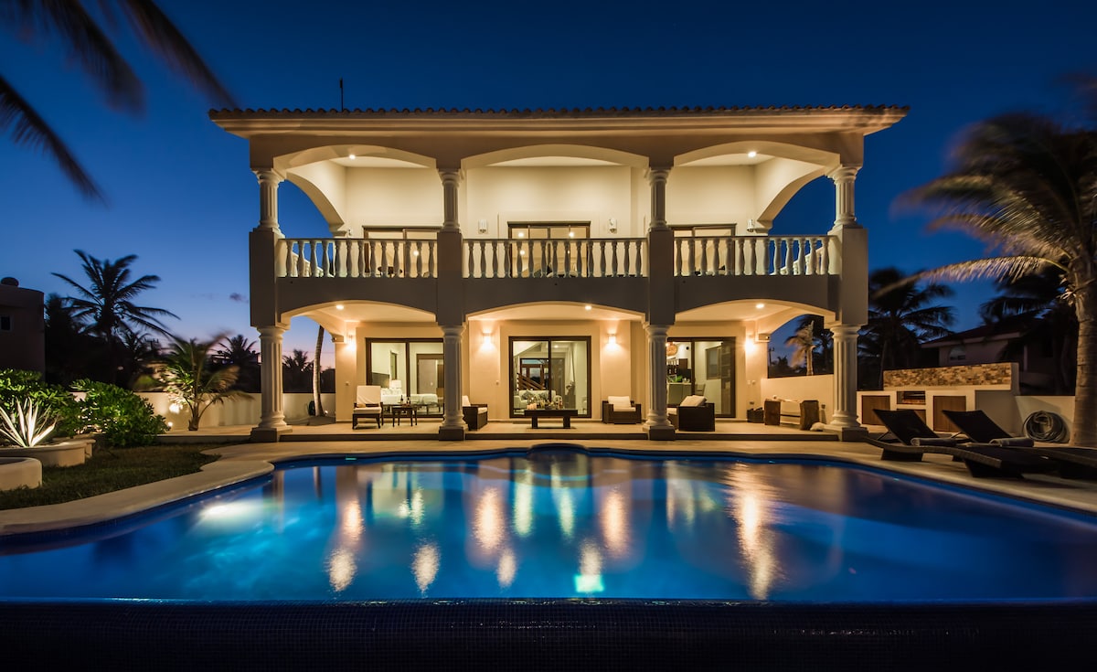 The villa is showcased at twilight, with illuminated architecture highlighting the spacious terrace and expansive windows. A large infinity pool reflects soft lighting, surrounded by palm trees and comfortable lounge chairs, creating an inviting outdoor space for relaxation.