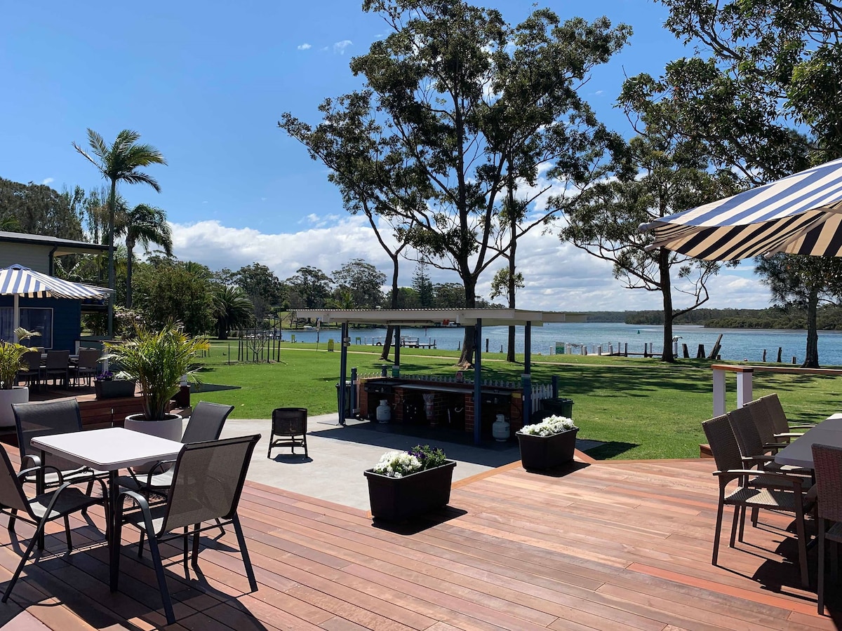 A spacious deck features a dining area with a table and chairs, surrounded by potted plants. The scene overlooks a serene inlet with palm trees in the background, providing a calm outdoor setting for relaxation and social gatherings.