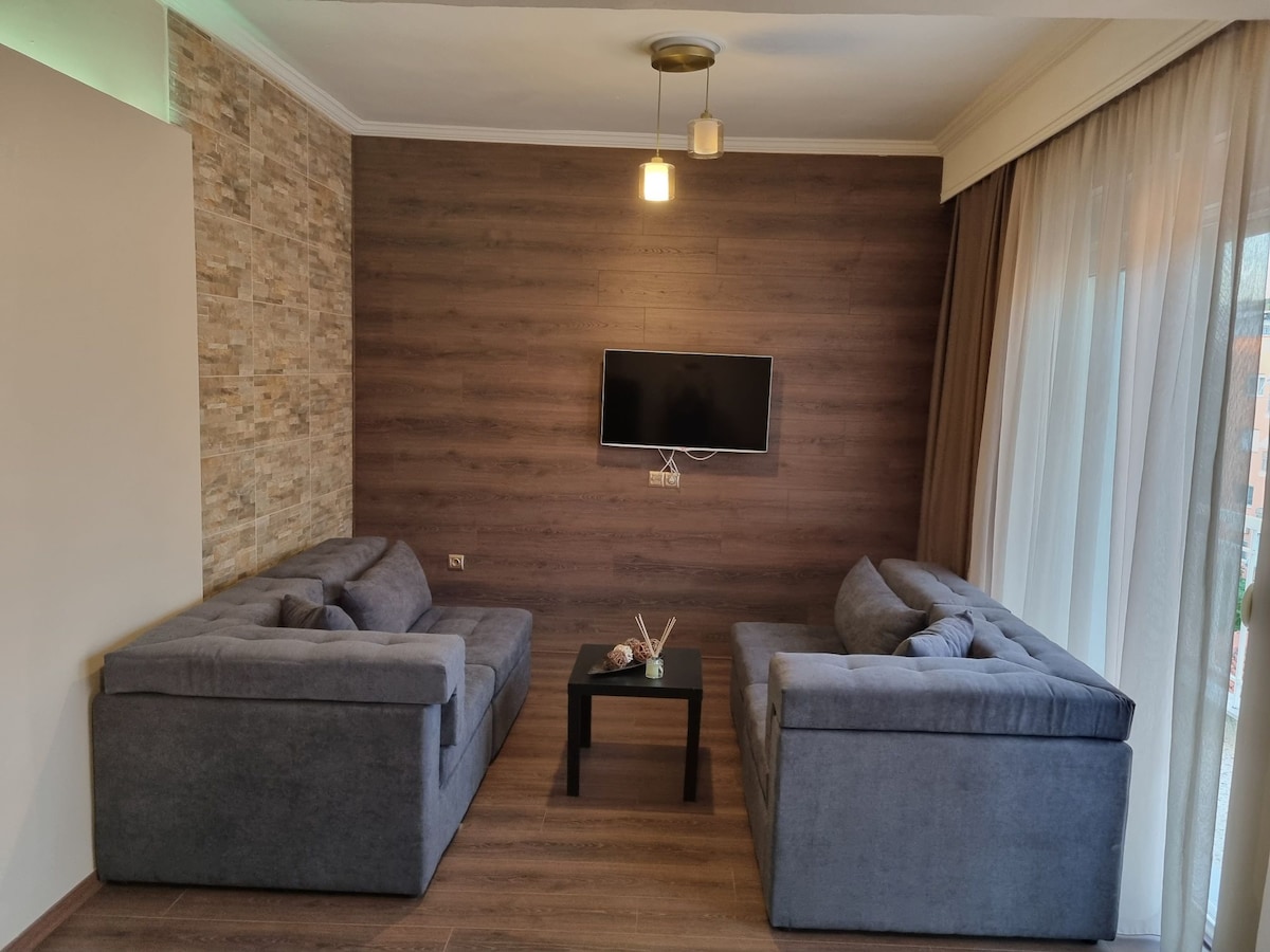 A modern living area is featured with two spacious sofas arranged opposite each other. A small black coffee table is situated between them, and a flat-screen television is mounted on the wooden wall. Large windows bring natural light into the space.